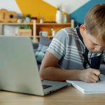 Young boy studies at a laptop and notebook.